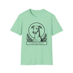 Holy Orders | Greyhound T-Shirt flat lay on sage green shirt featuring a stained glass style illustration of a Greyhound depicted as a holy figure with a golden halo and the text 'Holy Orders.'