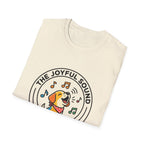 The Joyful Sound of Barking T-shirt folded view, natural tan shirt showing the circular badge graphic of a barking cartoon dog with musical notes and the text 'The Joyful Sound of Barking.'