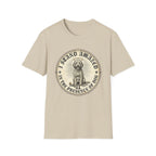 I Stand Amazed Golden Doodle T-Shirt flat lay in sand, featuring a circular vintage-style graphic of a fluffy golden doodle with a halo surrounded by rays of light, with the text "I Stand Amazed in the Presence of Dog" and a small human figure kneeling before the dog.