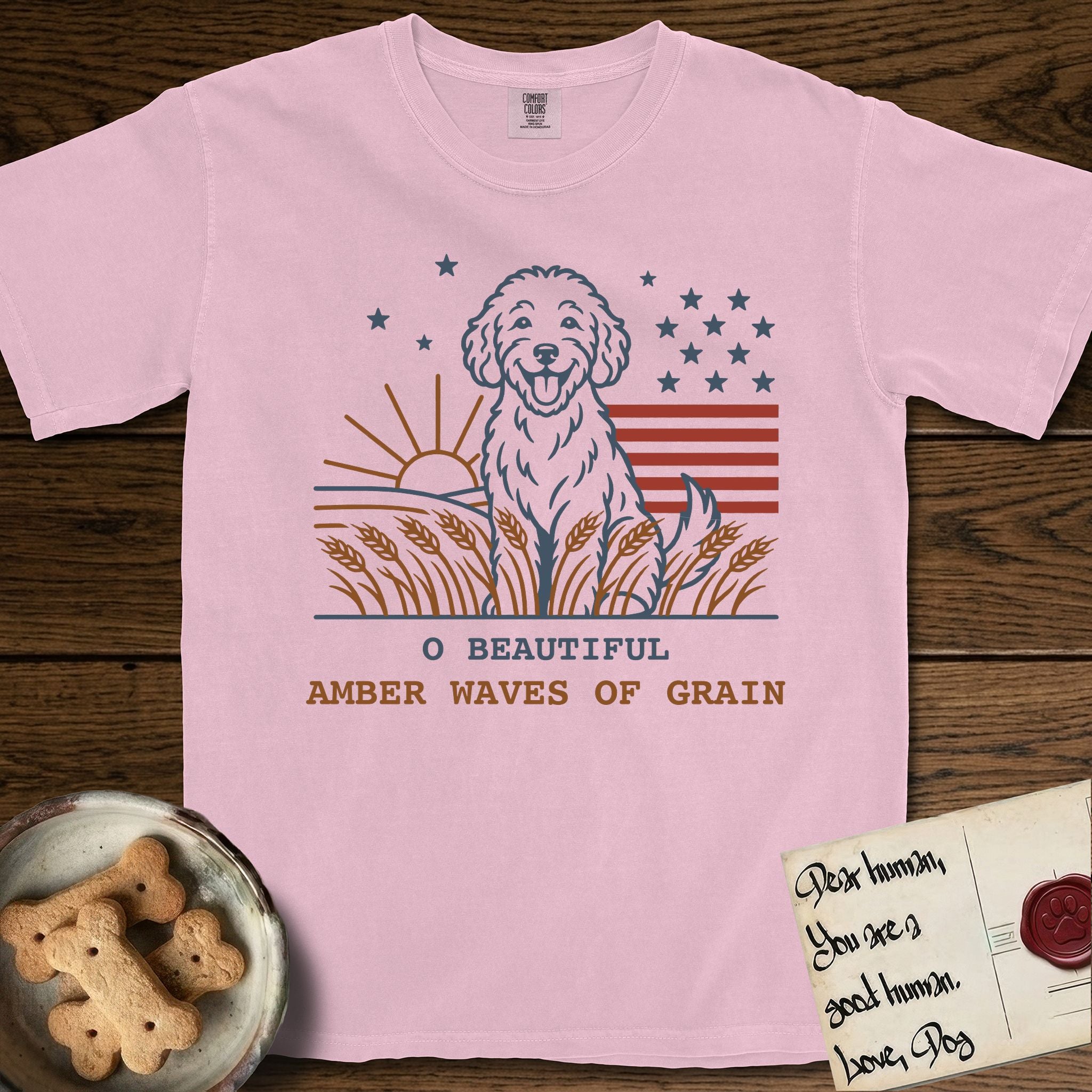 Amber Waves of Grain T-Shirt flat lay in Blossom, featuring a faded vintage-Americana line-art Goldendoodle sitting in a wheat field with a rising sun, scattered stars, and red flag stripes, above “O Beautiful / Amber Waves of Grain” lettering.