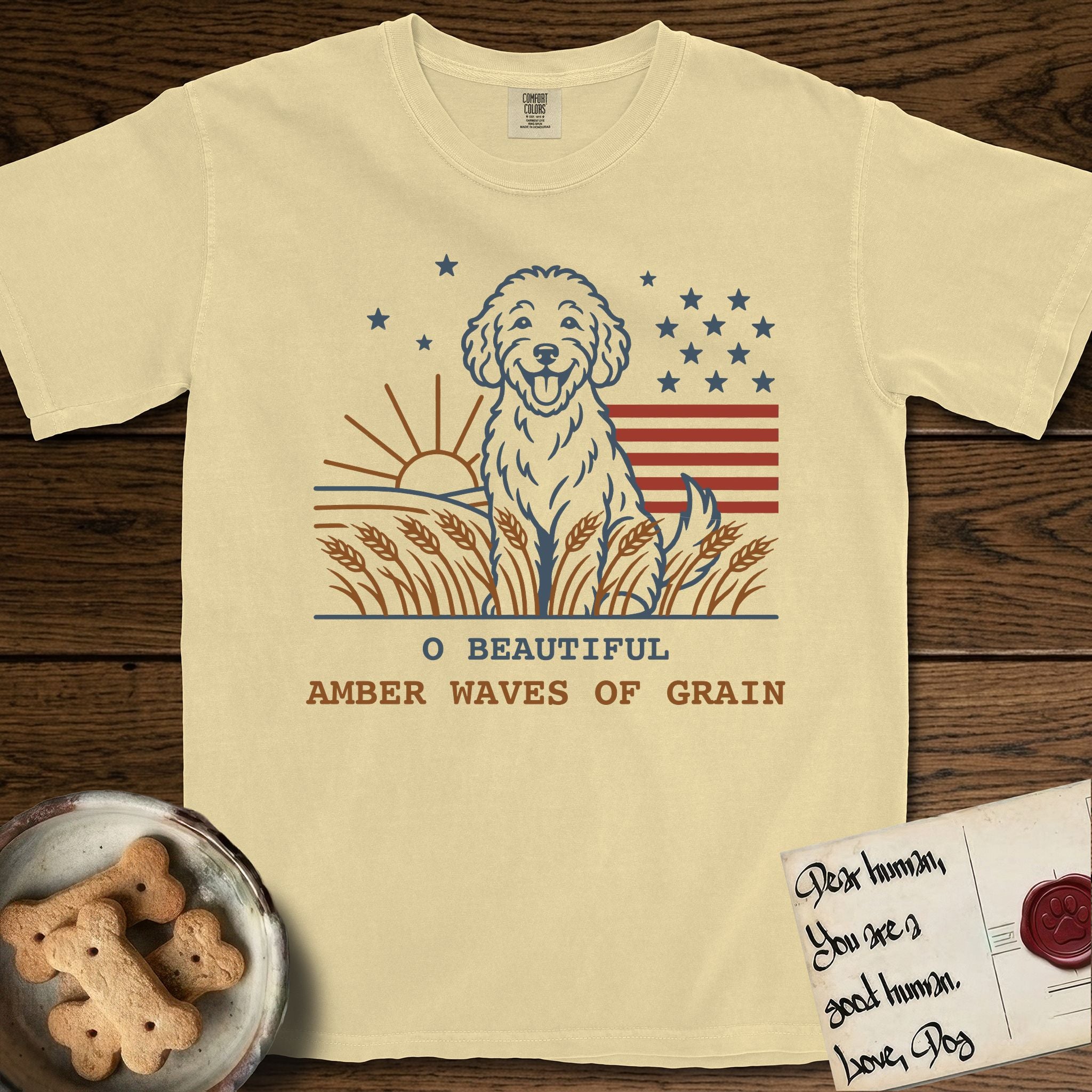 Amber Waves of Grain T-Shirt flat lay in Butter, featuring a faded vintage-Americana line-art Goldendoodle sitting in a wheat field with a rising sun, scattered stars, and red flag stripes, above “O Beautiful / Amber Waves of Grain” lettering.
