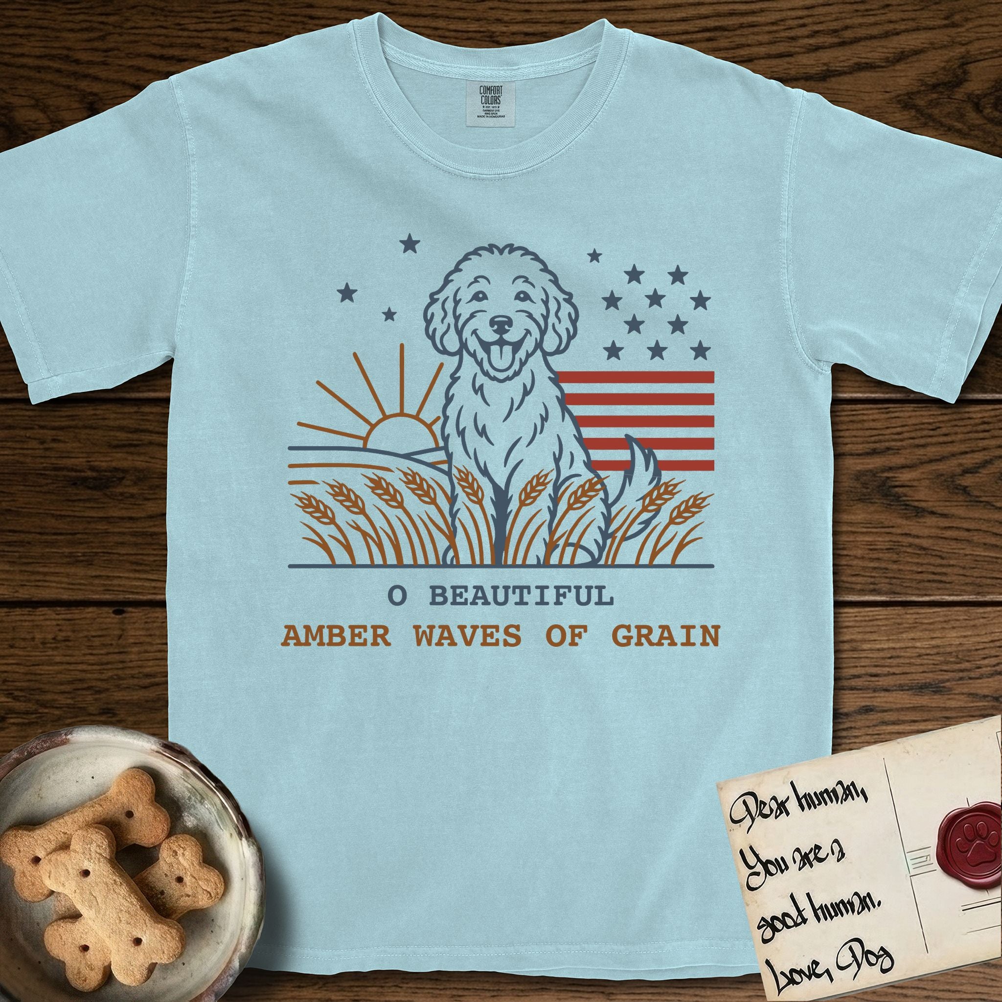 Amber Waves of Grain T-Shirt flat lay in Chambray, featuring a faded vintage-Americana line-art Goldendoodle sitting in a wheat field with a rising sun, scattered stars, and red flag stripes, above “O Beautiful / Amber Waves of Grain” lettering.
