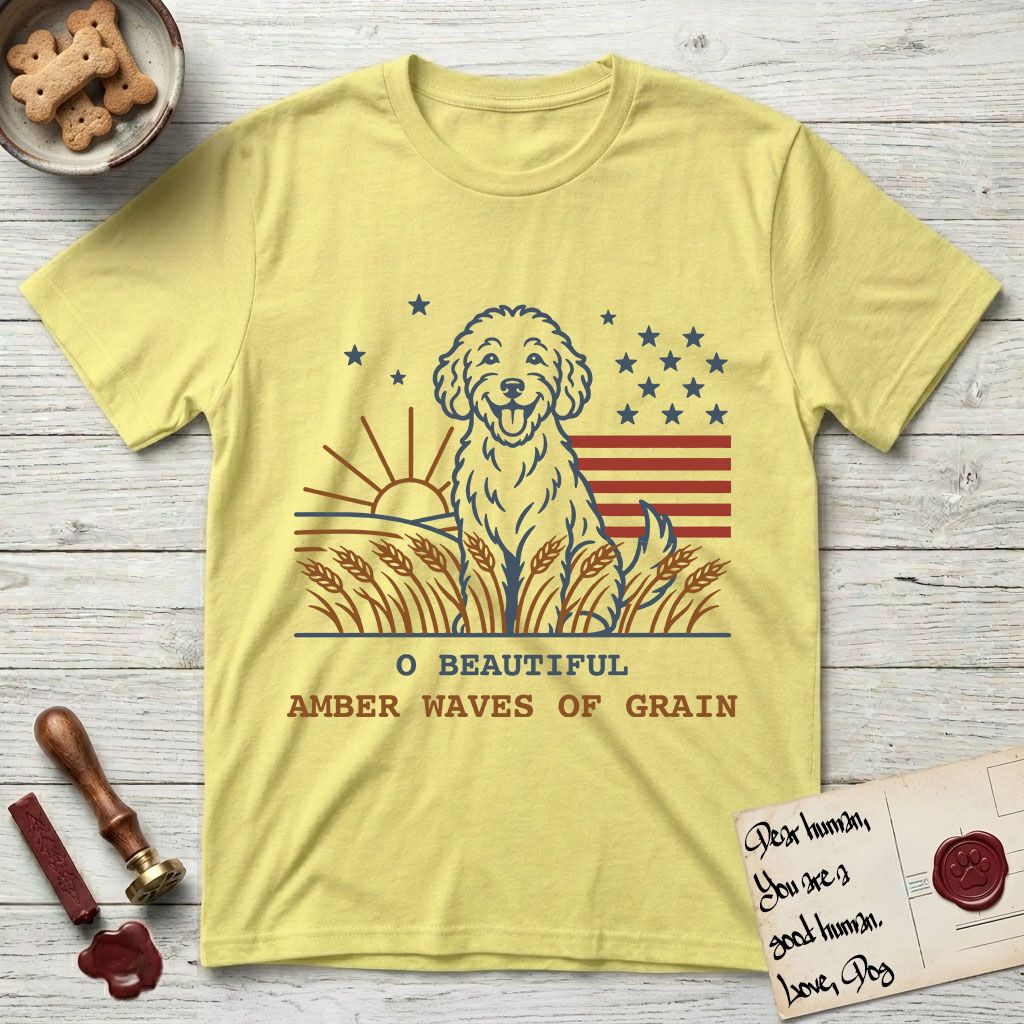 Amber Waves of Grain T-Shirt flat lay in Cornsilk, featuring a faded vintage-Americana line-art Goldendoodle sitting in a wheat field with a rising sun, scattered stars, and red flag stripes, above “O Beautiful / Amber Waves of Grain” lettering.