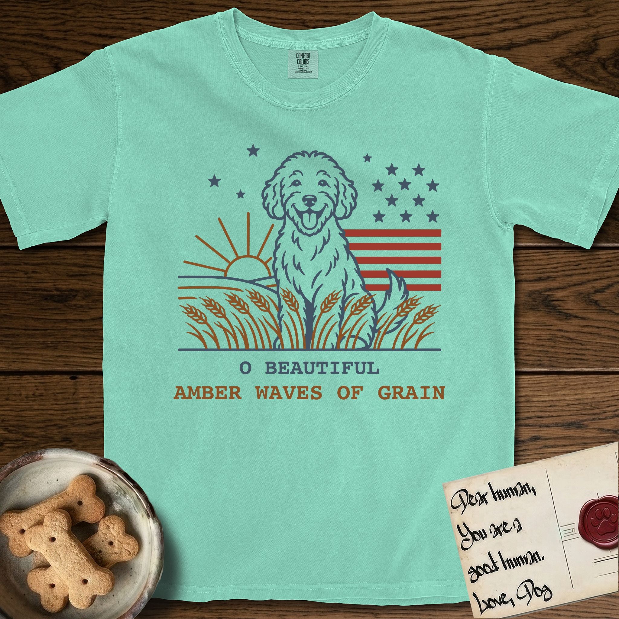 Amber Waves of Grain T-Shirt flat lay in Island Reef, featuring a faded vintage-Americana line-art Goldendoodle sitting in a wheat field with a rising sun, scattered stars, and red flag stripes, above “O Beautiful / Amber Waves of Grain” lettering.