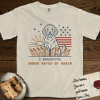 Amber Waves of Grain T-Shirt flat lay in Ivory, featuring a faded vintage-Americana line-art Goldendoodle sitting in a wheat field with a rising sun, scattered stars, and red flag stripes, above “O Beautiful / Amber Waves of Grain” lettering.