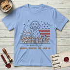 Amber Waves of Grain T-Shirt flat lay in Light Blue, featuring a faded vintage-Americana line-art Goldendoodle sitting in a wheat field with a rising sun, scattered stars, and red flag stripes, above “O Beautiful / Amber Waves of Grain” lettering.