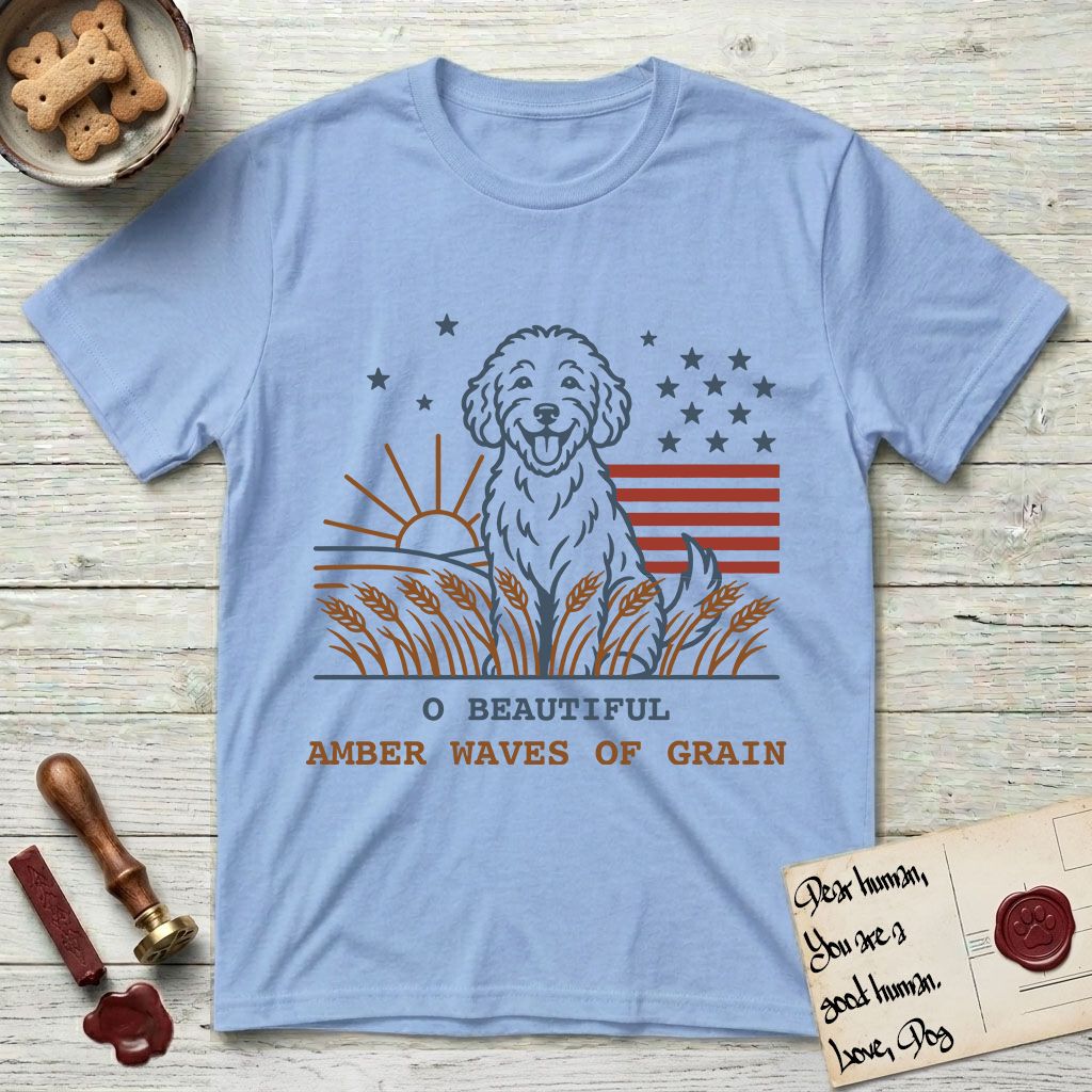 Amber Waves of Grain T-Shirt flat lay in Light Blue, featuring a faded vintage-Americana line-art Goldendoodle sitting in a wheat field with a rising sun, scattered stars, and red flag stripes, above “O Beautiful / Amber Waves of Grain” lettering.