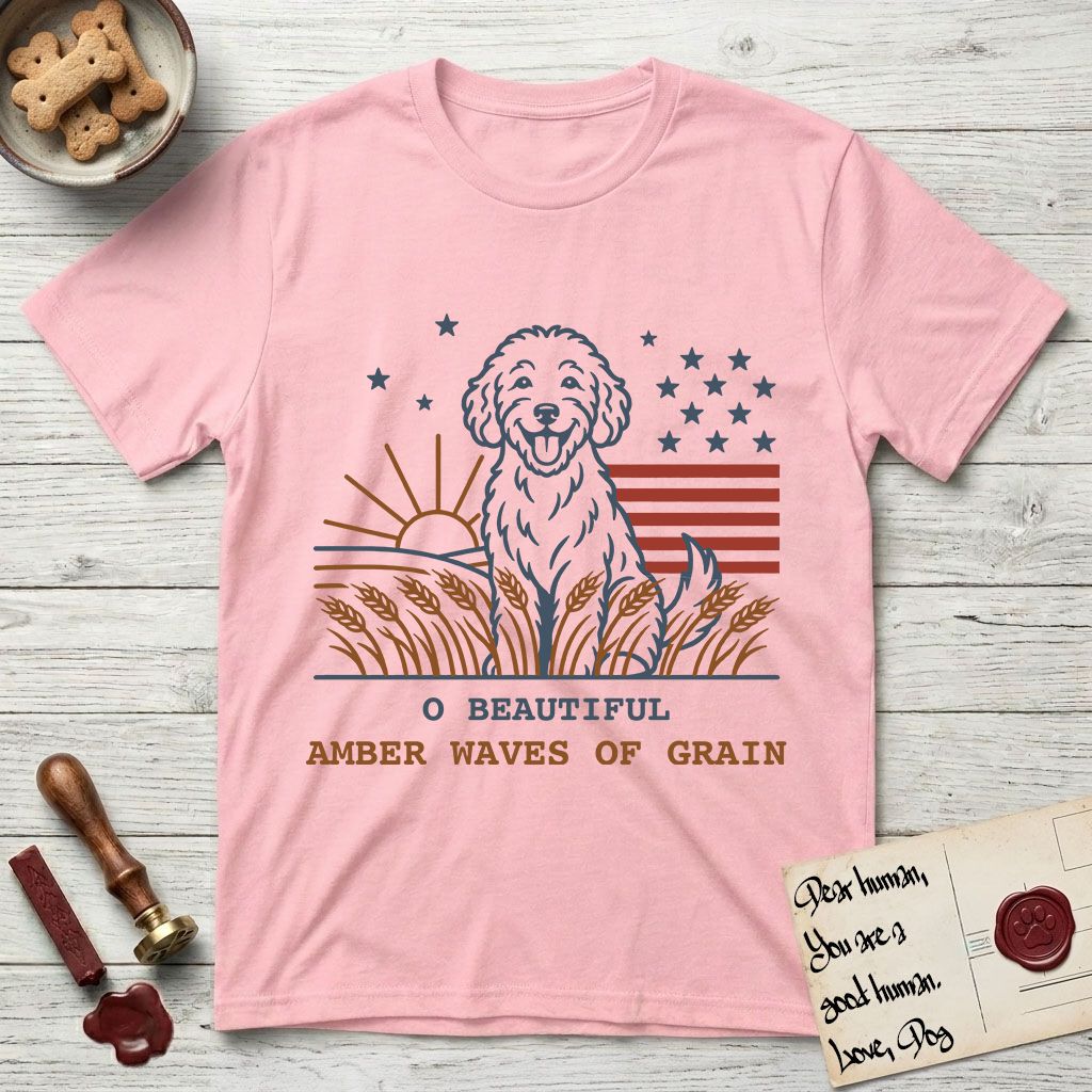 Amber Waves of Grain T-Shirt flat lay in Light Pink, featuring a faded vintage-Americana line-art Goldendoodle sitting in a wheat field with a rising sun, scattered stars, and red flag stripes, above “O Beautiful / Amber Waves of Grain” lettering.