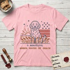 Amber Waves of Grain T-Shirt flat lay in Light Pink, featuring a faded vintage-Americana line-art Goldendoodle sitting in a wheat field with a rising sun, scattered stars, and red flag stripes, above “O Beautiful / Amber Waves of Grain” lettering.
