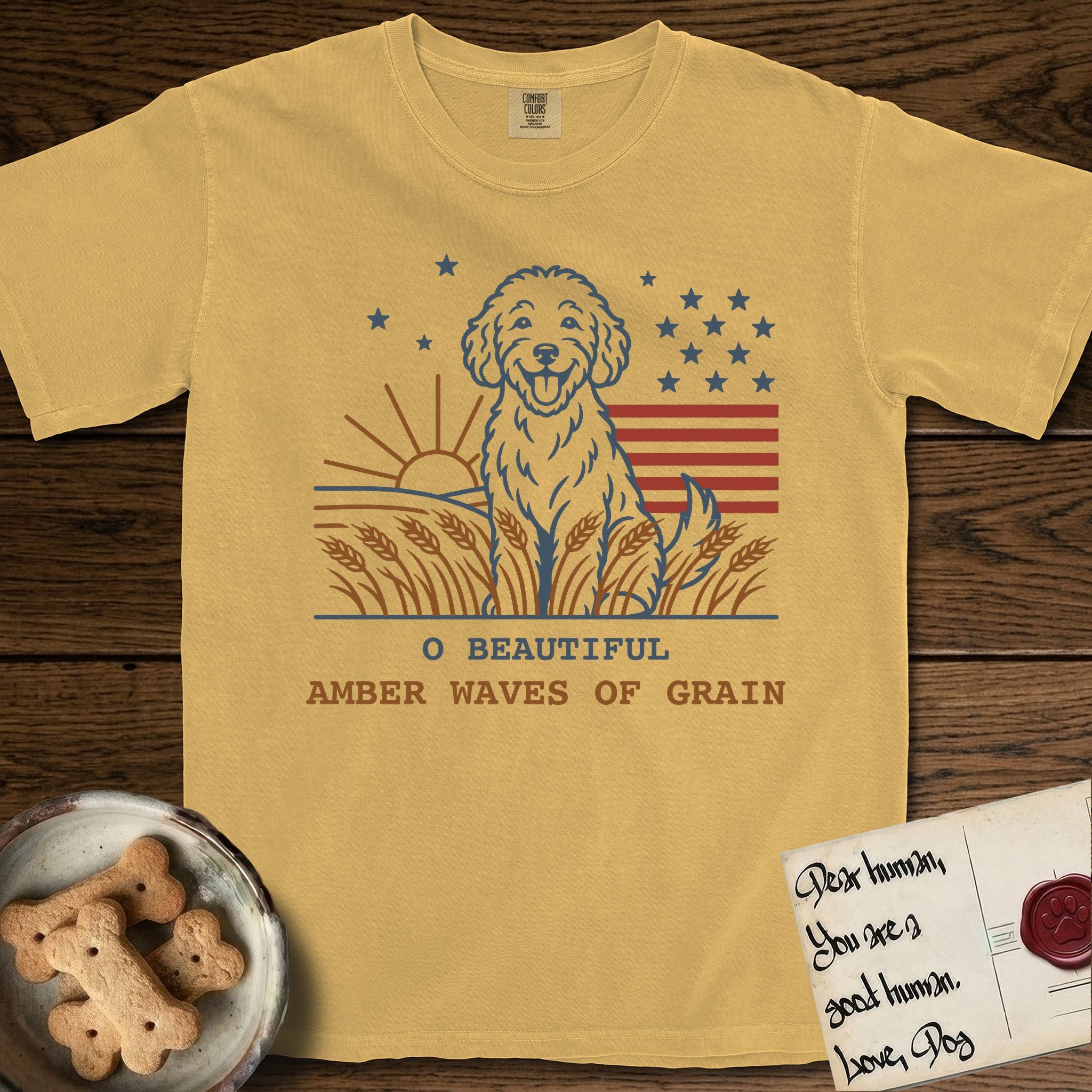 Amber Waves of Grain T-Shirt flat lay in Mustard, featuring a faded vintage-Americana line-art Goldendoodle sitting in a wheat field with a rising sun, scattered stars, and red flag stripes, above “O Beautiful / Amber Waves of Grain” lettering.