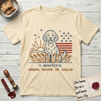 Amber Waves of Grain T-Shirt flat lay in Natural, featuring a faded vintage-Americana line-art Goldendoodle sitting in a wheat field with a rising sun, scattered stars, and red flag stripes, above “O Beautiful / Amber Waves of Grain” lettering.