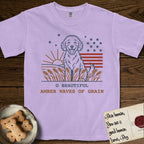 Amber Waves of Grain T-Shirt flat lay in Orchid, featuring a faded vintage-Americana line-art Goldendoodle sitting in a wheat field with a rising sun, scattered stars, and red flag stripes, above “O Beautiful / Amber Waves of Grain” lettering.