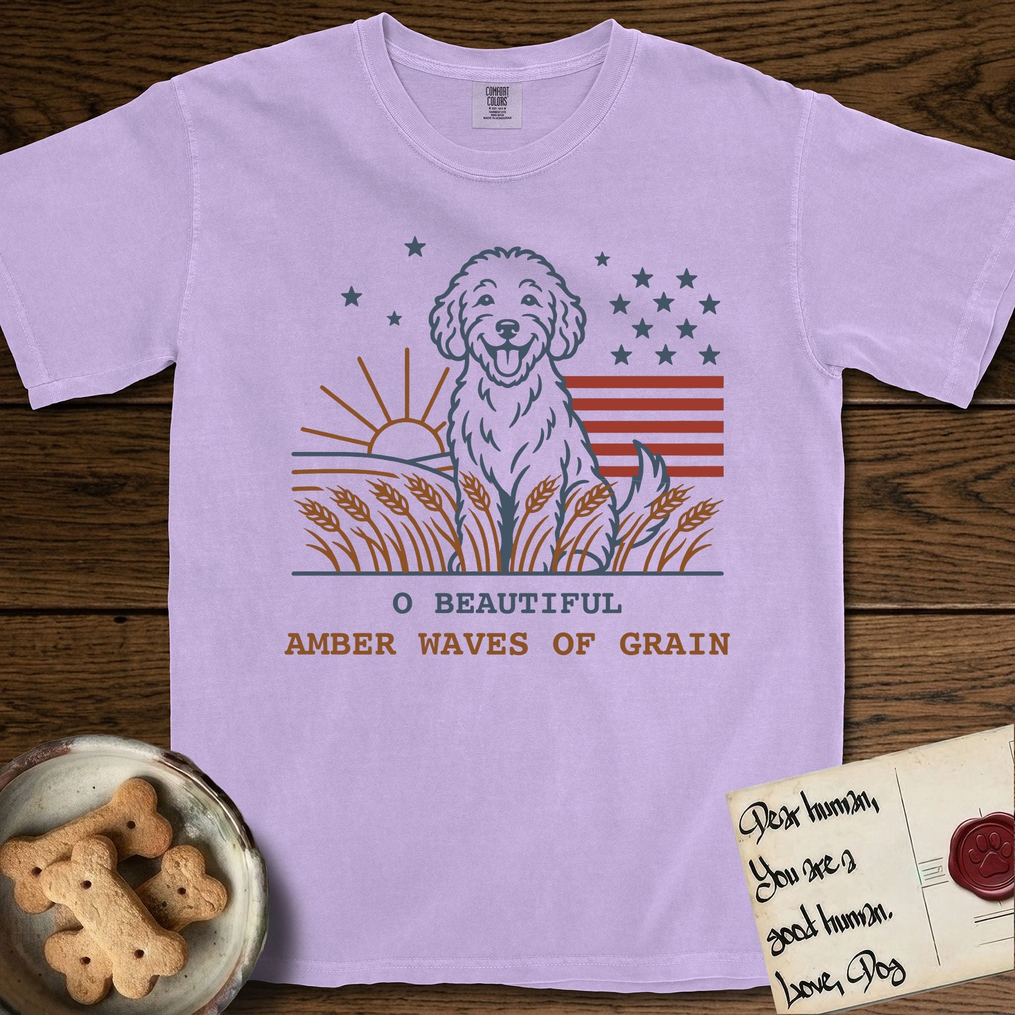 Amber Waves of Grain T-Shirt flat lay in Orchid, featuring a faded vintage-Americana line-art Goldendoodle sitting in a wheat field with a rising sun, scattered stars, and red flag stripes, above “O Beautiful / Amber Waves of Grain” lettering.