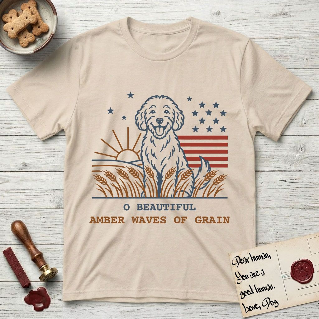 Amber Waves of Grain T-Shirt flat lay in Sand, featuring a faded vintage-Americana line-art Goldendoodle sitting in a wheat field with a rising sun, scattered stars, and red flag stripes, above “O Beautiful / Amber Waves of Grain” lettering.