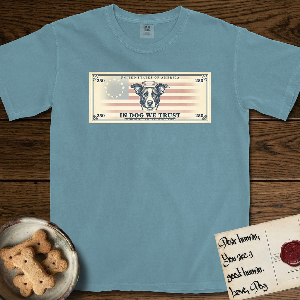 In Dog We Trust Bill T-Shirt flat lay in Ice Blue, featuring a vintage dollar-bill design with a haloed mixed-breed Dog at center, a Betsy Ross flag in the background, and bold currency-style "In Dog We Trust" typography.
