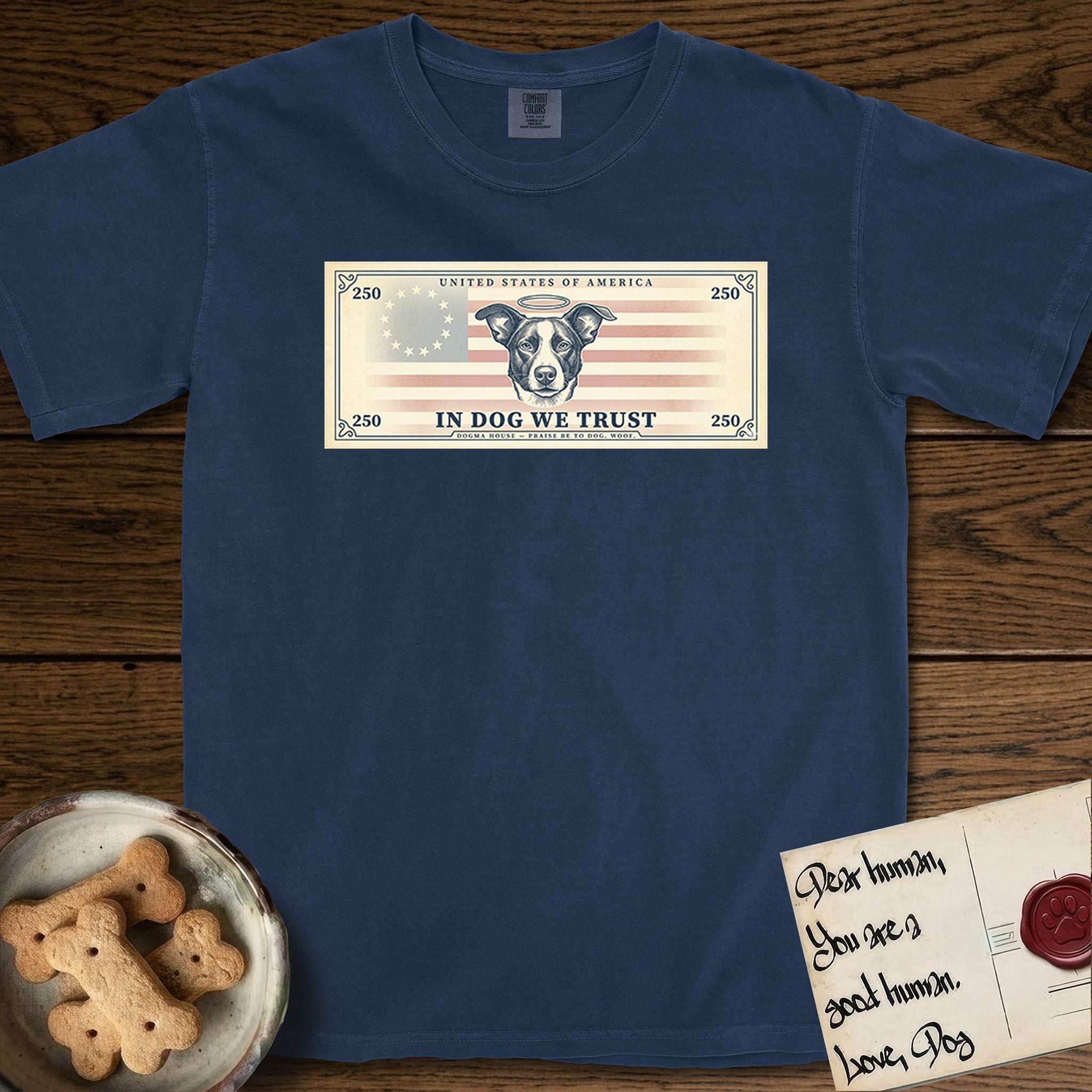 In Dog We Trust Bill T-Shirt flat lay in True Navy, featuring a vintage dollar-bill design with a haloed mixed-breed Dog at center, a Betsy Ross flag in the background, and bold currency-style "In Dog We Trust" typography.