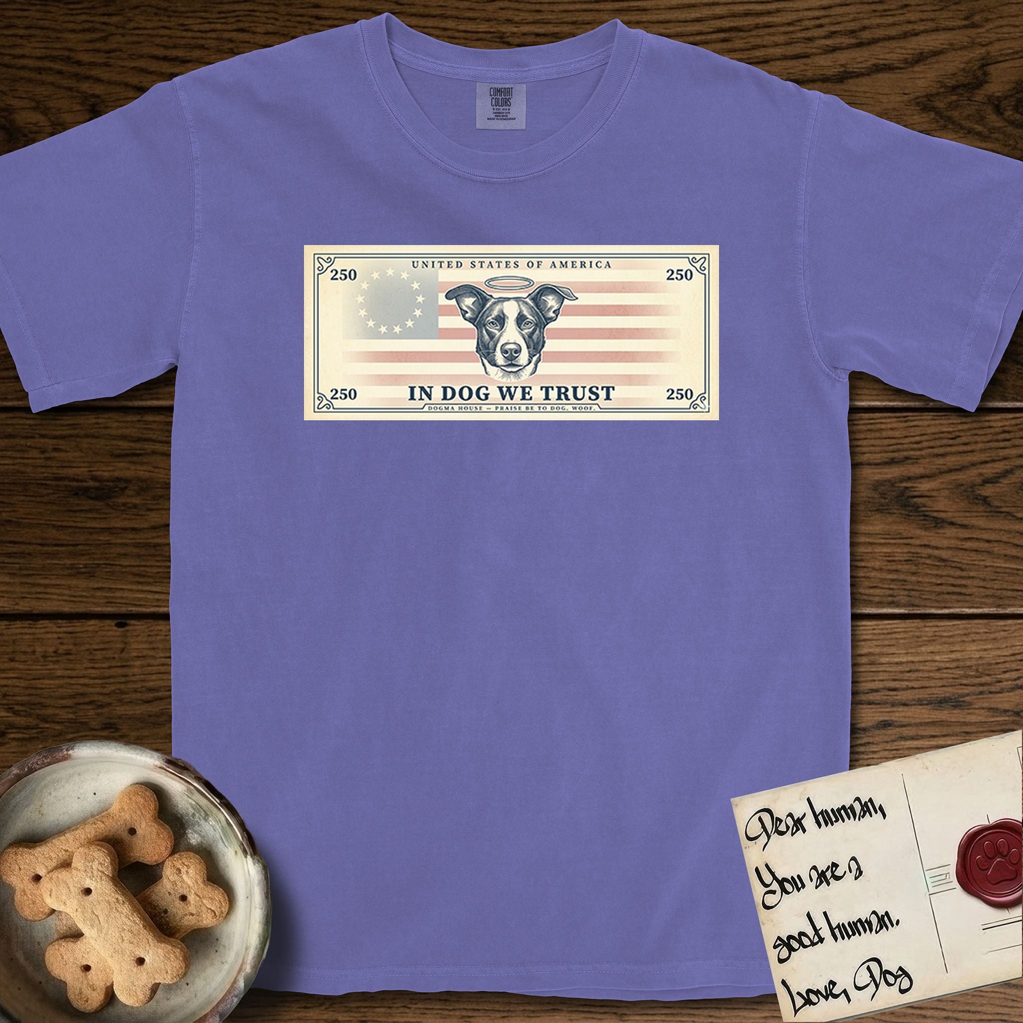 In Dog We Trust Bill T-Shirt flat lay in Violet, featuring a vintage dollar-bill design with a haloed mixed-breed Dog at center, a Betsy Ross flag in the background, and bold currency-style "In Dog We Trust" typography.