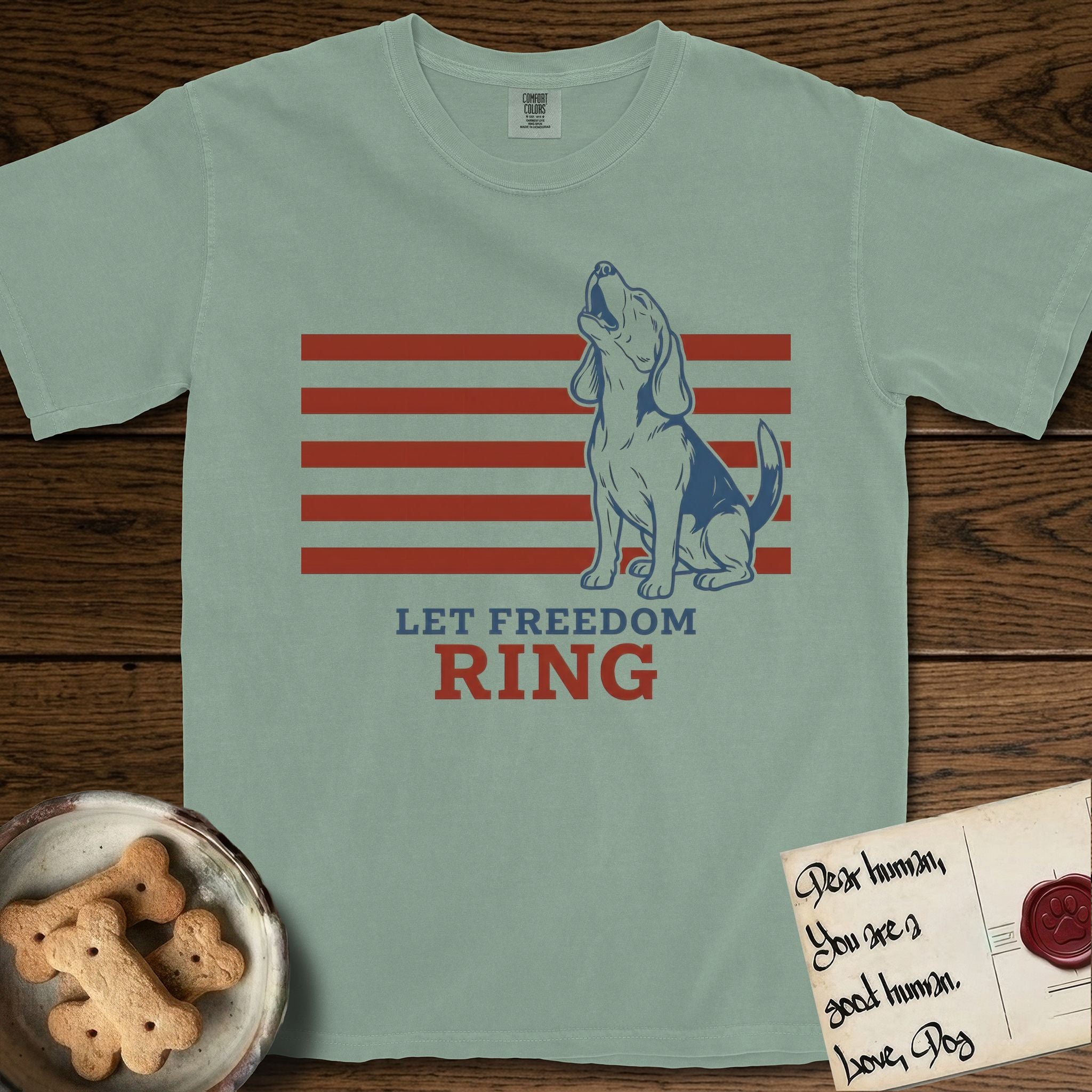 Let Freedom Ring T-Shirt flat lay in Bay, featuring a faded vintage-Americana line-art Beagle sitting and baying with her head thrown back in front of red flag stripes, above “Let Freedom Ring” lettering.