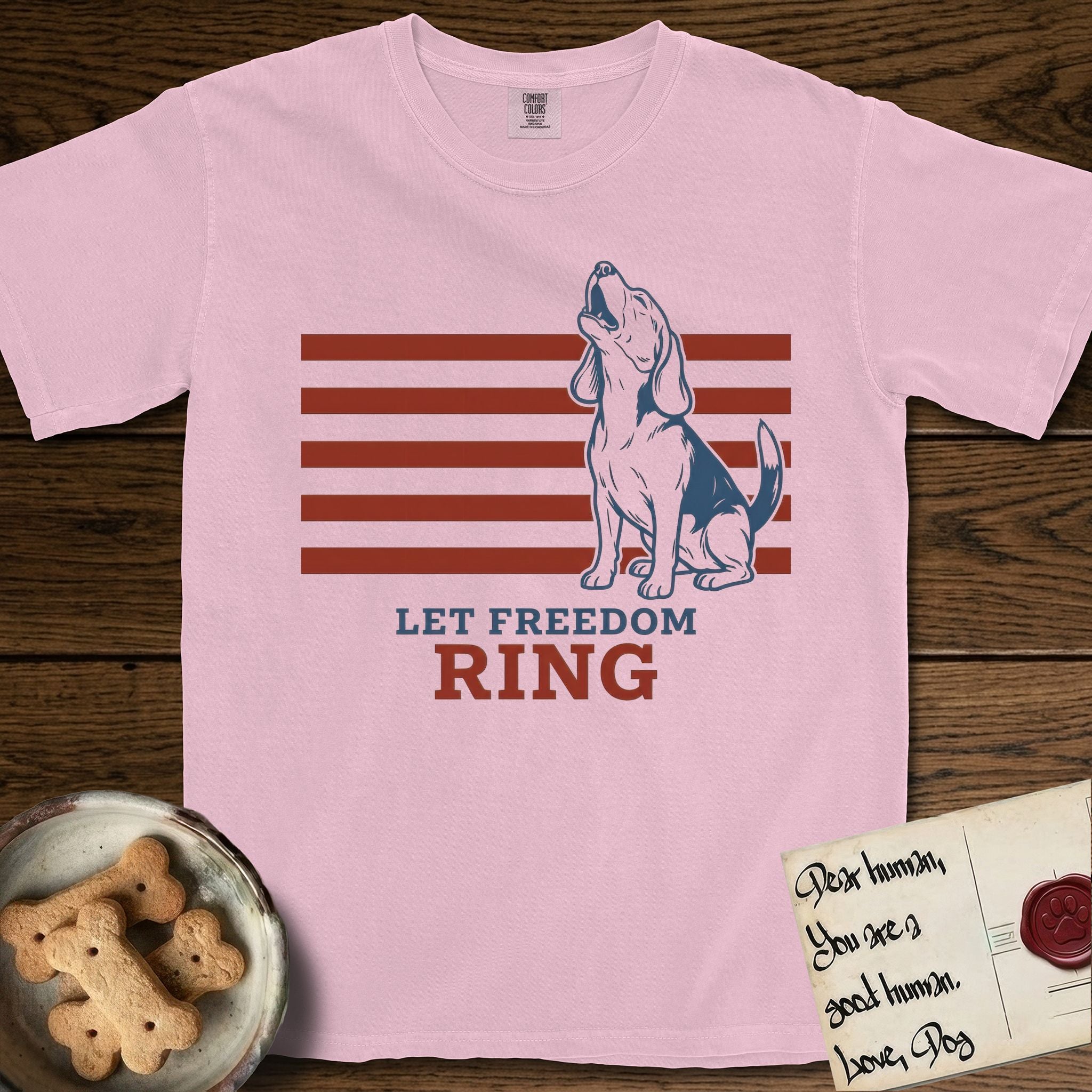 Let Freedom Ring T-Shirt flat lay in Blossom, featuring a faded vintage-Americana line-art Beagle sitting and baying with her head thrown back in front of red flag stripes, above “Let Freedom Ring” lettering.