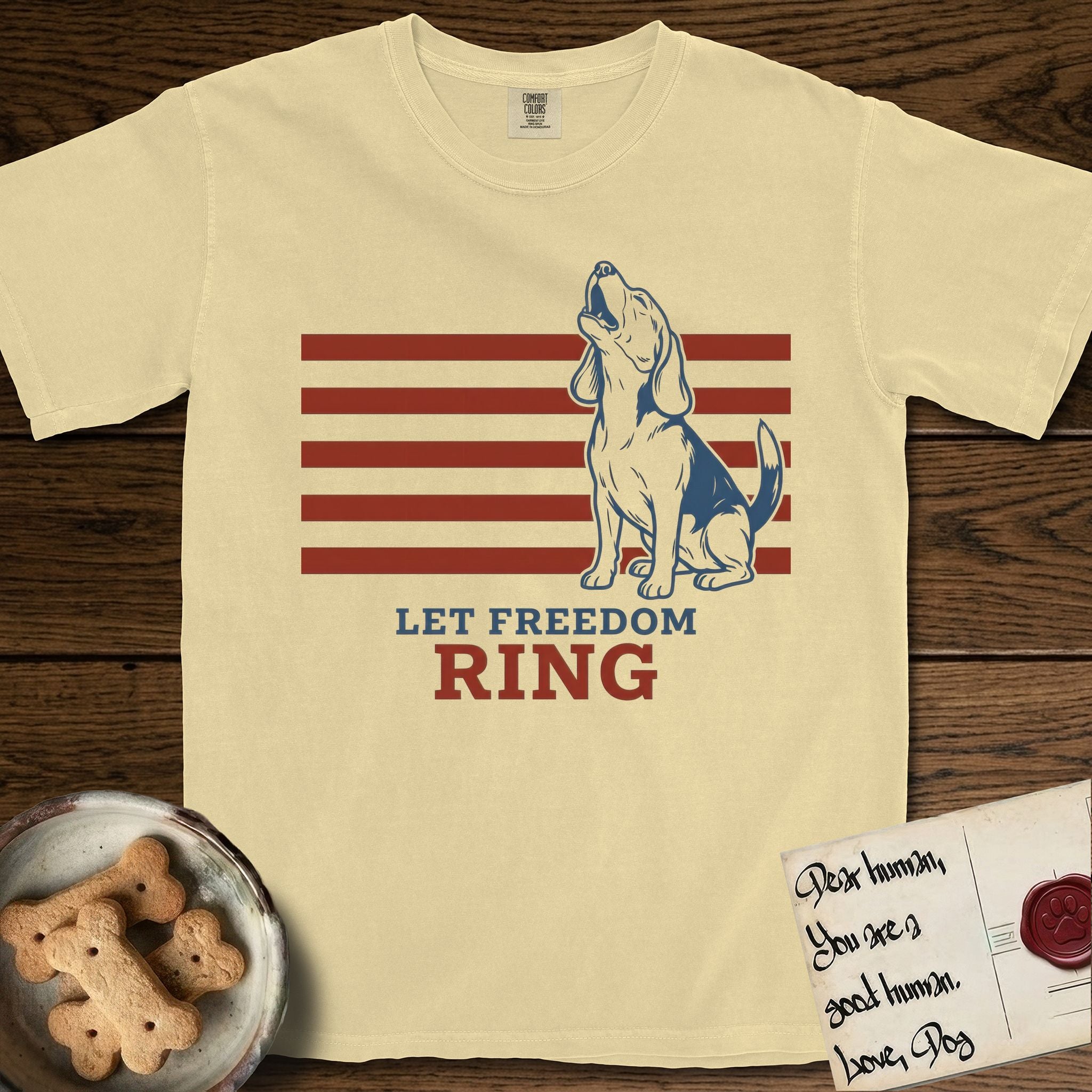 Let Freedom Ring T-Shirt flat lay in Butter, featuring a faded vintage-Americana line-art Beagle sitting and baying with her head thrown back in front of red flag stripes, above “Let Freedom Ring” lettering.