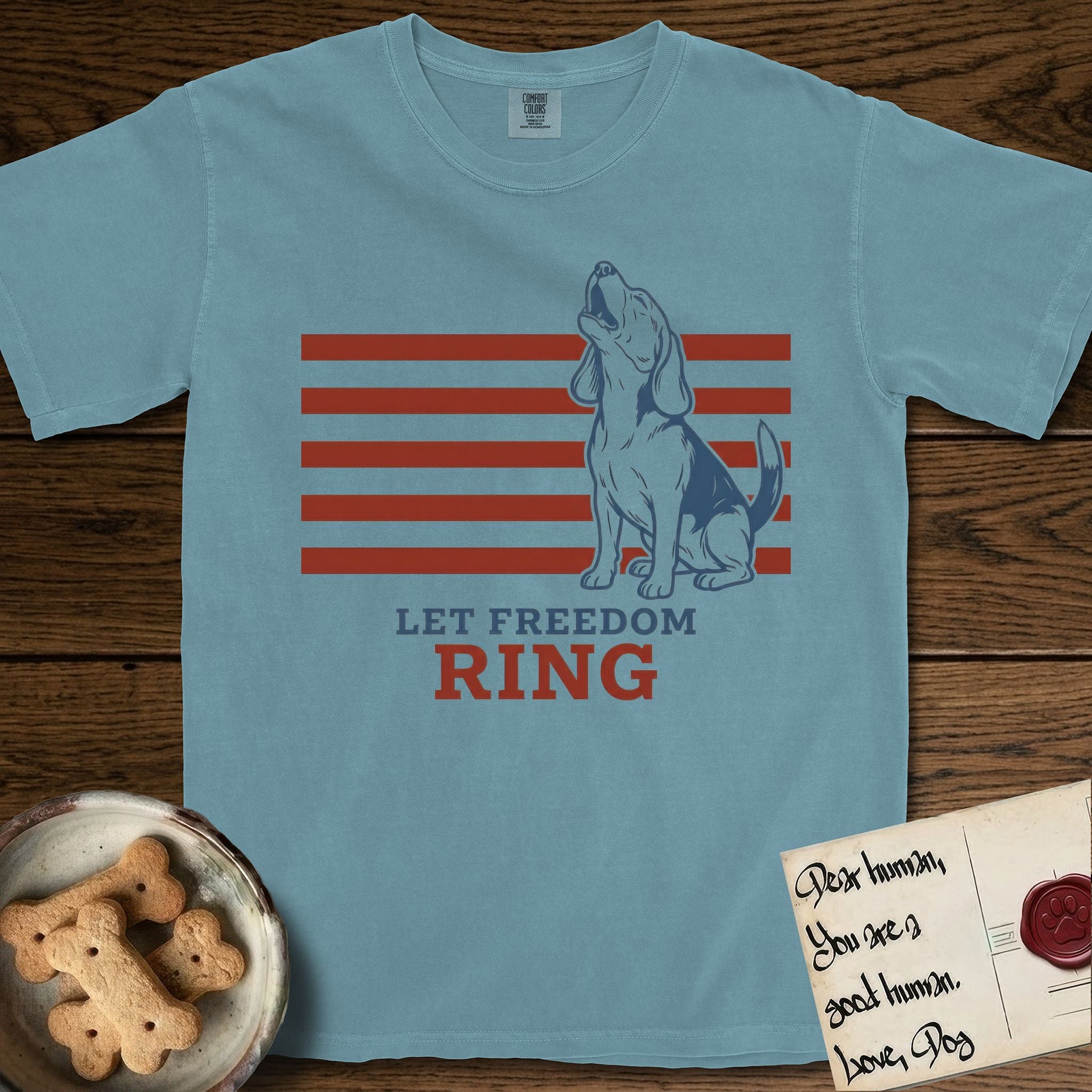 Let Freedom Ring T-Shirt flat lay in Ice Blue, featuring a faded vintage-Americana line-art Beagle sitting and baying with her head thrown back in front of red flag stripes, above “Let Freedom Ring” lettering.