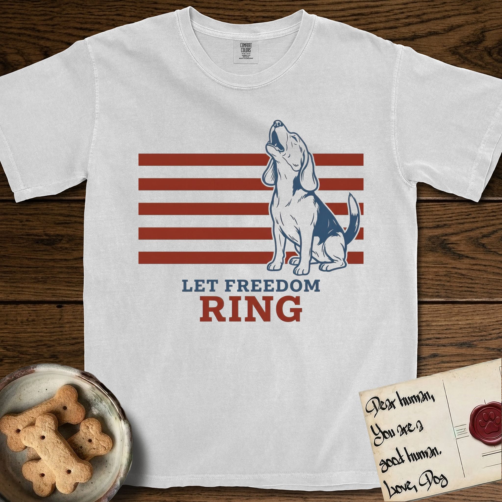 Let Freedom Ring T-Shirt flat lay in White, featuring a faded vintage-Americana line-art Beagle sitting and baying with her head thrown back in front of red flag stripes, above “Let Freedom Ring” lettering.