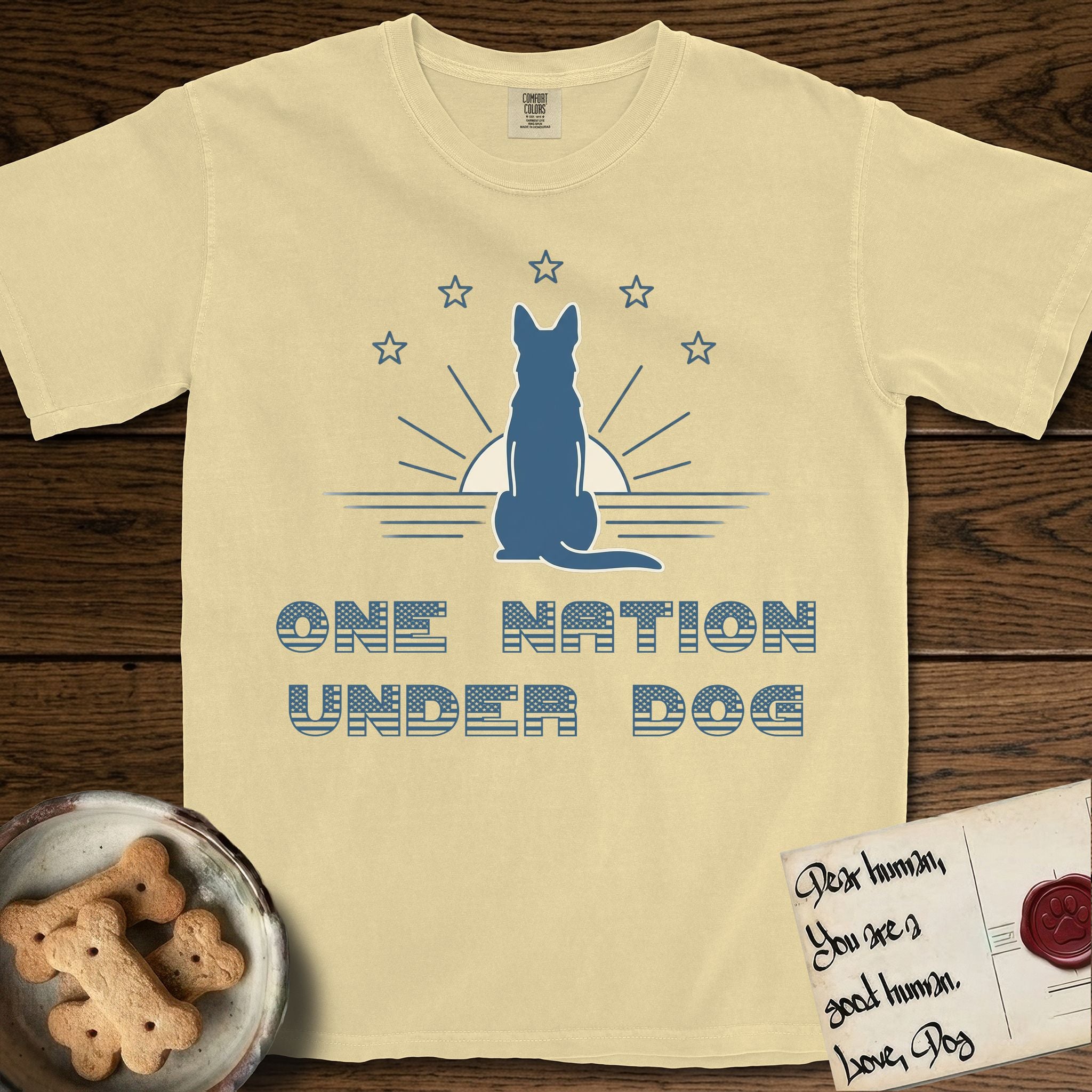 One Nation Under Dog T-Shirt flat lay in Butter, featuring a minimalist design with a Dog silhouette over a radiating sunburst, five stars, and bold "One Nation Under Dog" typography.