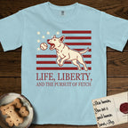 Life, Liberty, and the Pursuit of Fetch T-Shirt flat lay in Chambray, featuring a square flag-inspired design with a Bull Terrier leaping after a ball, cream stars, and bold "Life, Liberty, and the Pursuit of Fetch" serif typography.