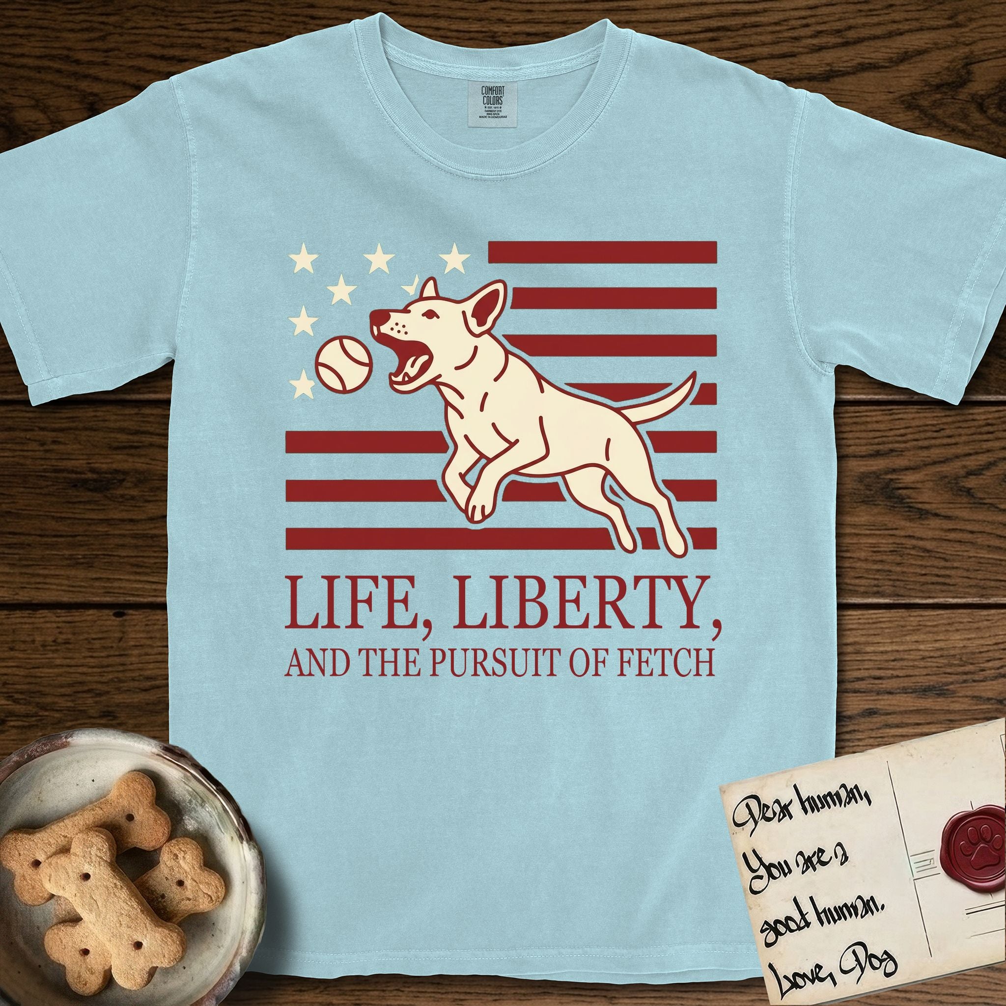 Life, Liberty, and the Pursuit of Fetch T-Shirt flat lay in Chambray, featuring a square flag-inspired design with a Bull Terrier leaping after a ball, cream stars, and bold "Life, Liberty, and the Pursuit of Fetch" serif typography.