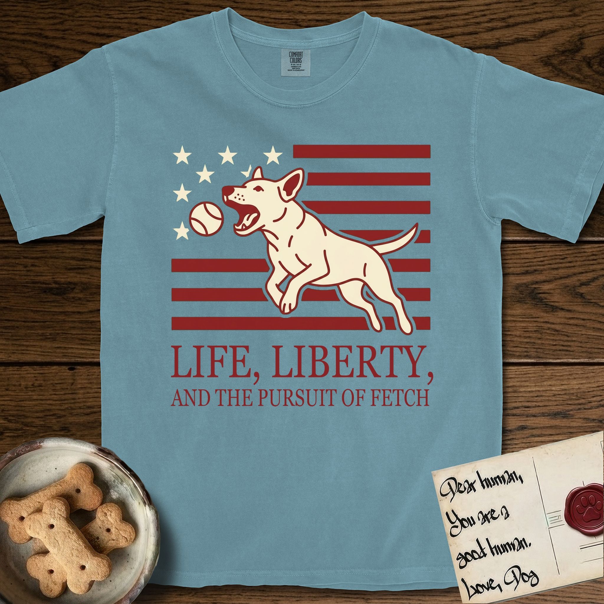 Life, Liberty, and the Pursuit of Fetch T-Shirt flat lay in Ice Blue, featuring a square flag-inspired design with a Bull Terrier leaping after a ball, cream stars, and bold "Life, Liberty, and the Pursuit of Fetch" serif typography.