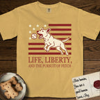 Life, Liberty, and the Pursuit of Fetch T-Shirt flat lay in Mustard, featuring a square flag-inspired design with a Bull Terrier leaping after a ball, cream stars, and bold "Life, Liberty, and the Pursuit of Fetch" serif typography.