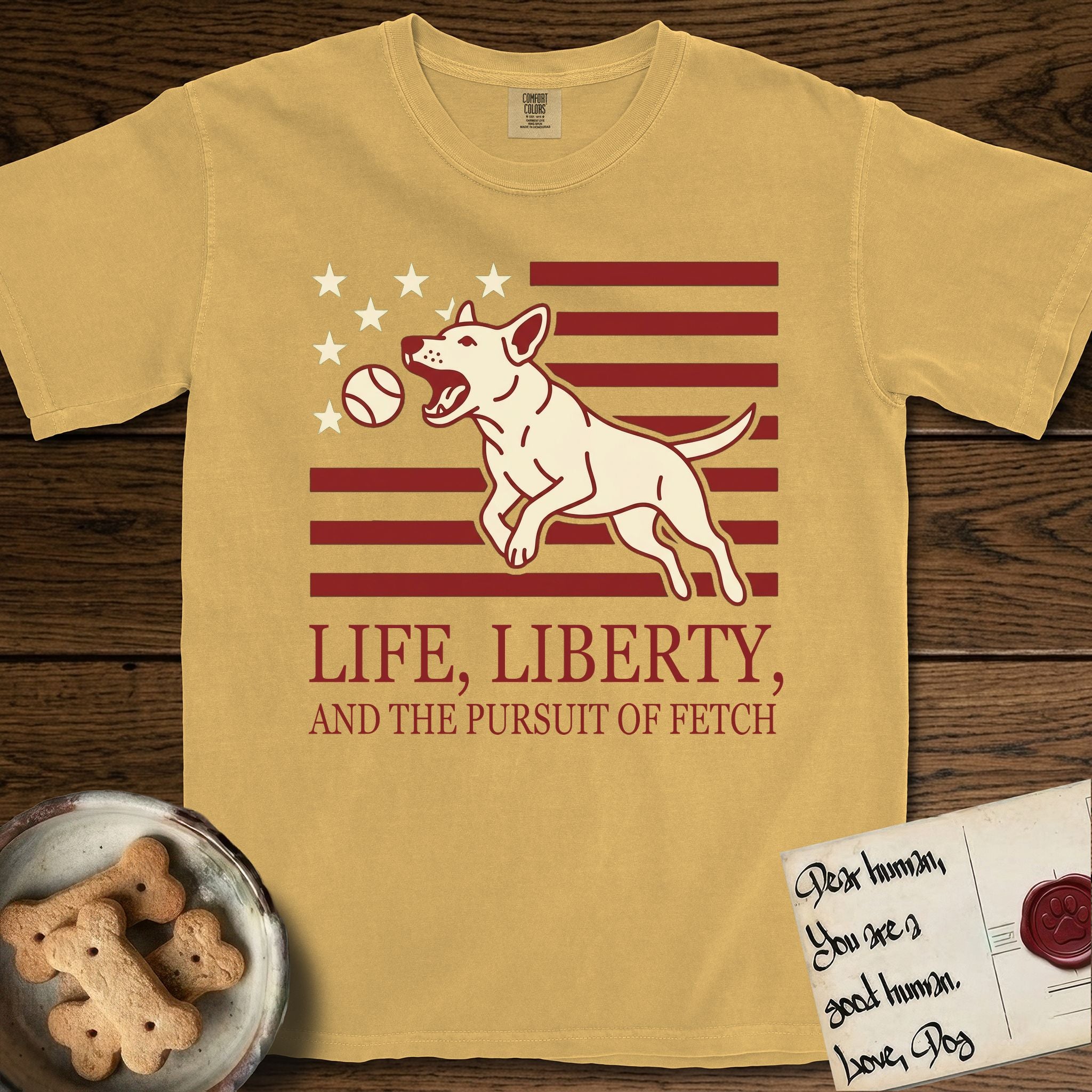 Life, Liberty, and the Pursuit of Fetch T-Shirt flat lay in Mustard, featuring a square flag-inspired design with a Bull Terrier leaping after a ball, cream stars, and bold "Life, Liberty, and the Pursuit of Fetch" serif typography.