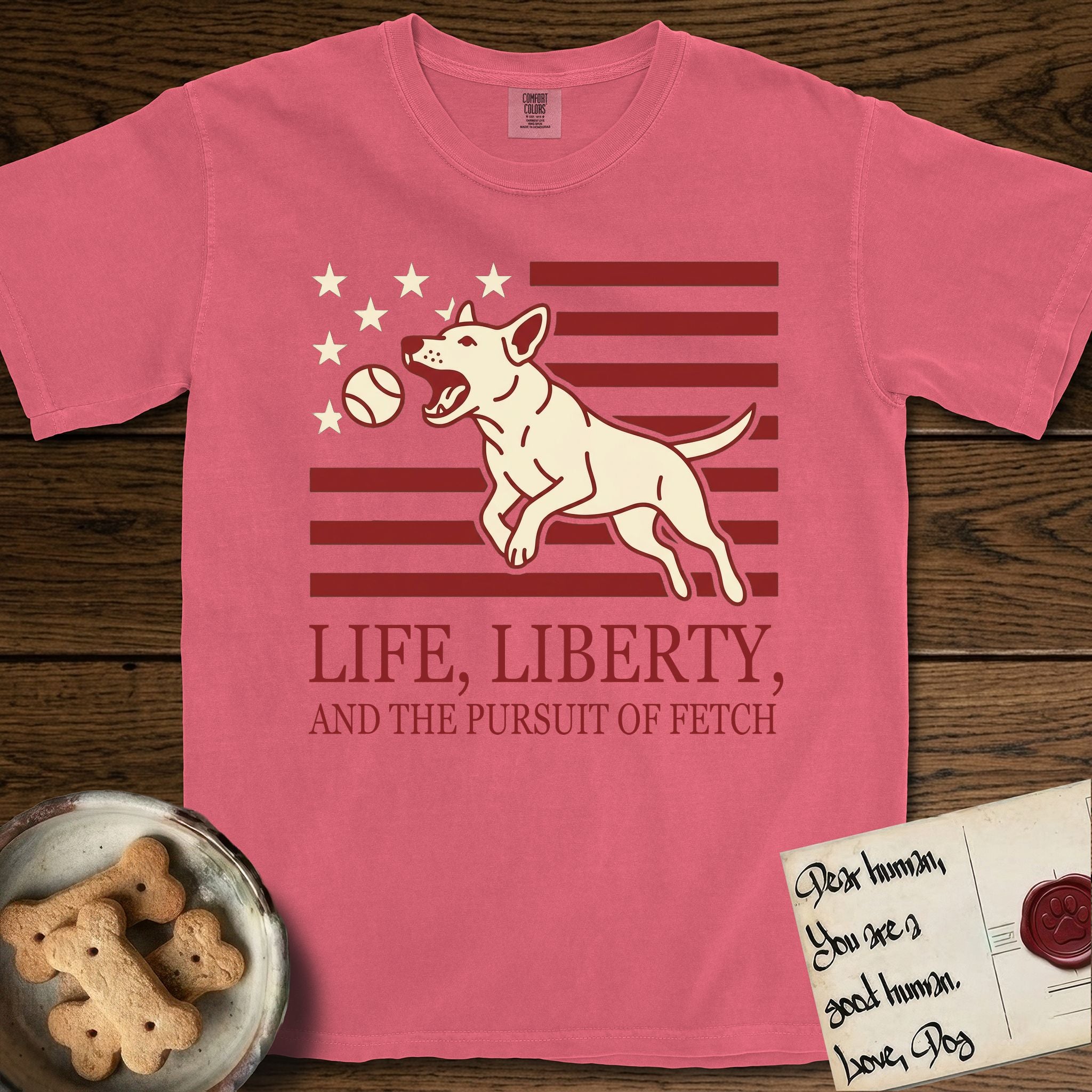 Life, Liberty, and the Pursuit of Fetch T-Shirt flat lay in Watermelon, featuring a square flag-inspired design with a Bull Terrier leaping after a ball, cream stars, and bold "Life, Liberty, and the Pursuit of Fetch" serif typography.