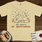 The Pursuit of Happiness T-Shirt flat lay in Butter, featuring a faded vintage-Americana line-art Samoyed rolling on his back in the grass with paws in the air under a sky of stars, above “The Pursuit of Happiness” lettering.