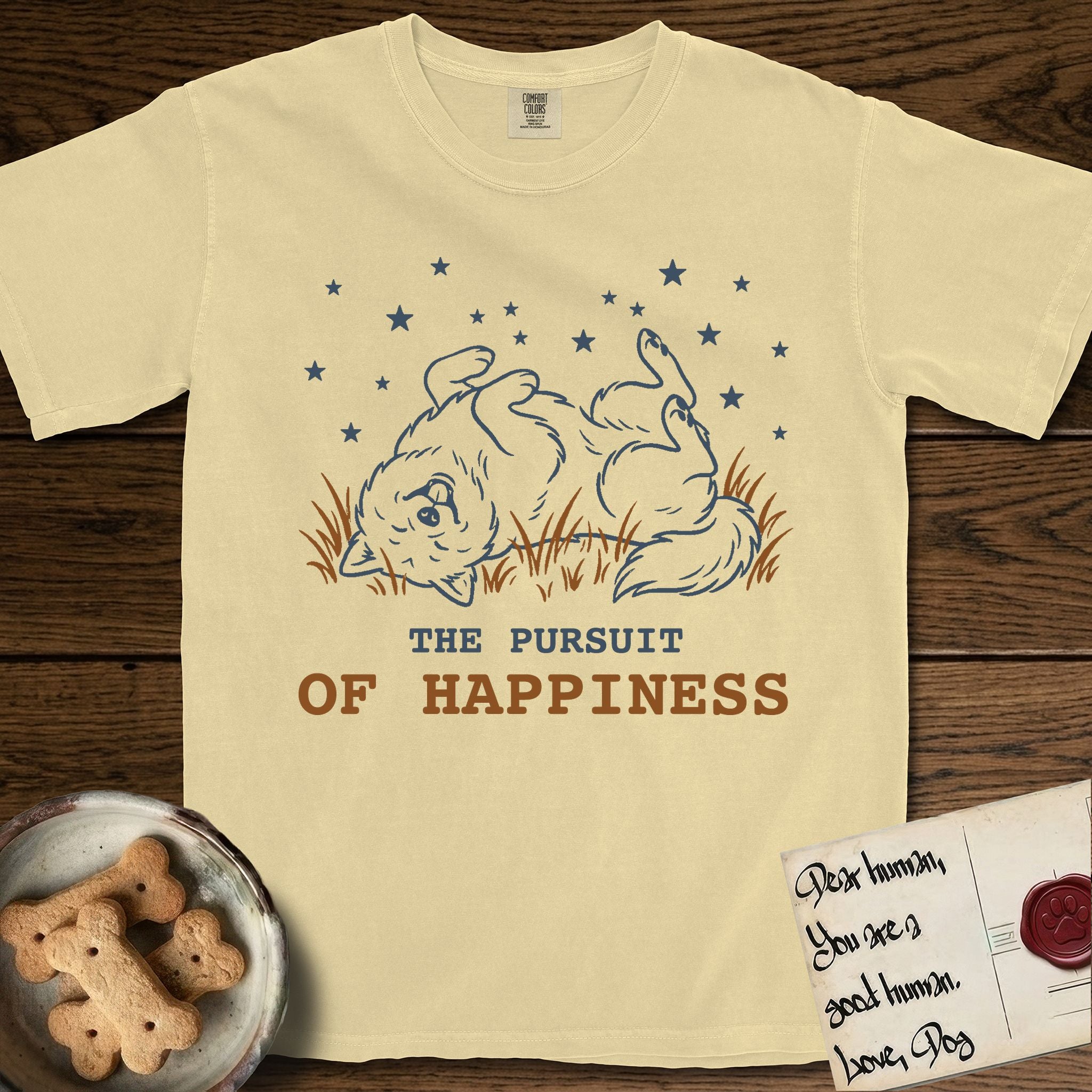 The Pursuit of Happiness T-Shirt flat lay in Butter, featuring a faded vintage-Americana line-art Samoyed rolling on his back in the grass with paws in the air under a sky of stars, above “The Pursuit of Happiness” lettering.