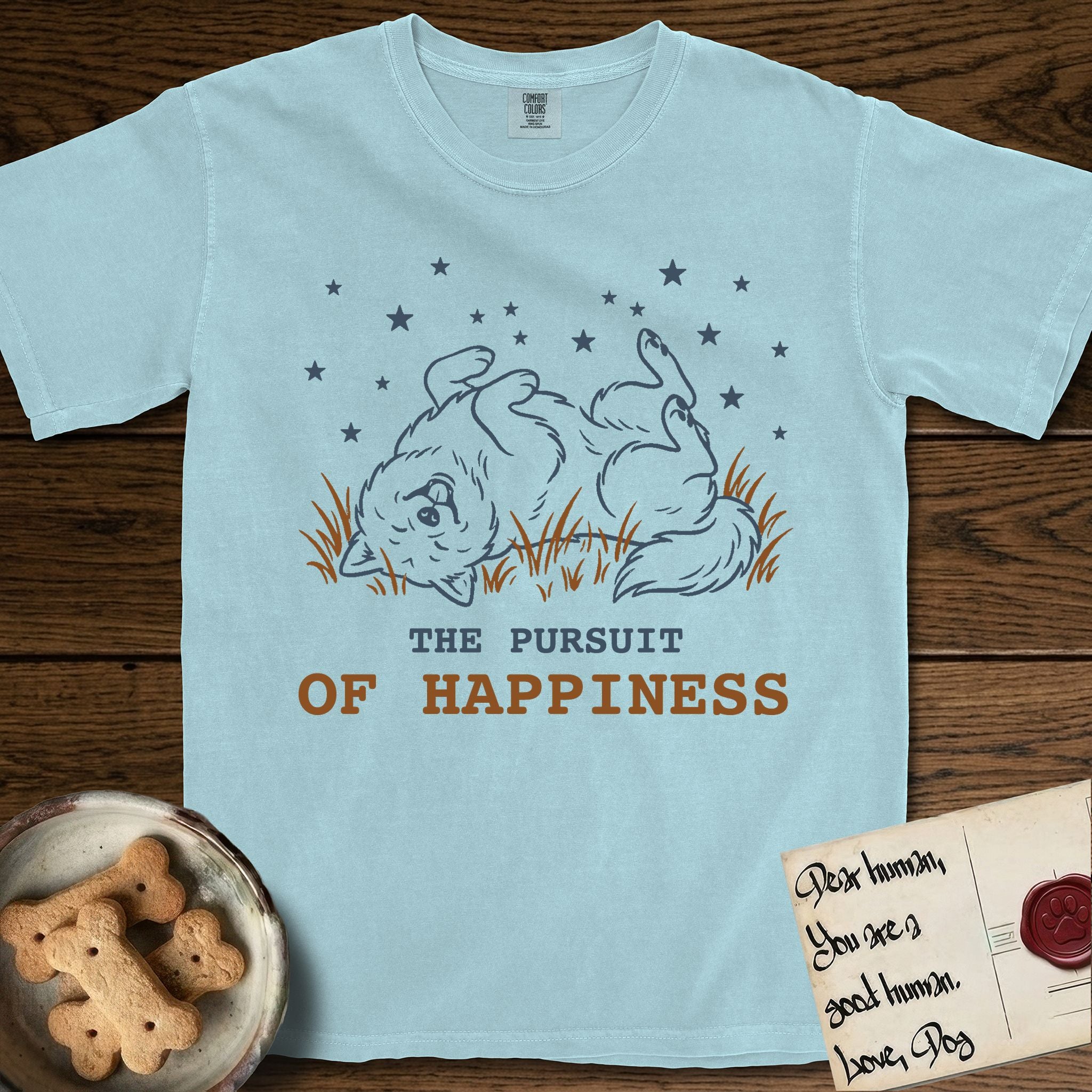 The Pursuit of Happiness T-Shirt flat lay in Chambray, featuring a faded vintage-Americana line-art Samoyed rolling on his back in the grass with paws in the air under a sky of stars, above “The Pursuit of Happiness” lettering.