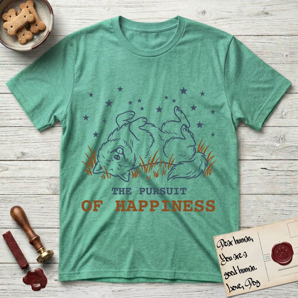The Pursuit of Happiness T-Shirt flat lay in Heather Irish Green, featuring a faded vintage-Americana line-art Samoyed rolling on his back in the grass with paws in the air under a sky of stars, above “The Pursuit of Happiness” lettering.