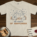The Pursuit of Happiness T-Shirt flat lay in Ivory, featuring a faded vintage-Americana line-art Samoyed rolling on his back in the grass with paws in the air under a sky of stars, above “The Pursuit of Happiness” lettering.