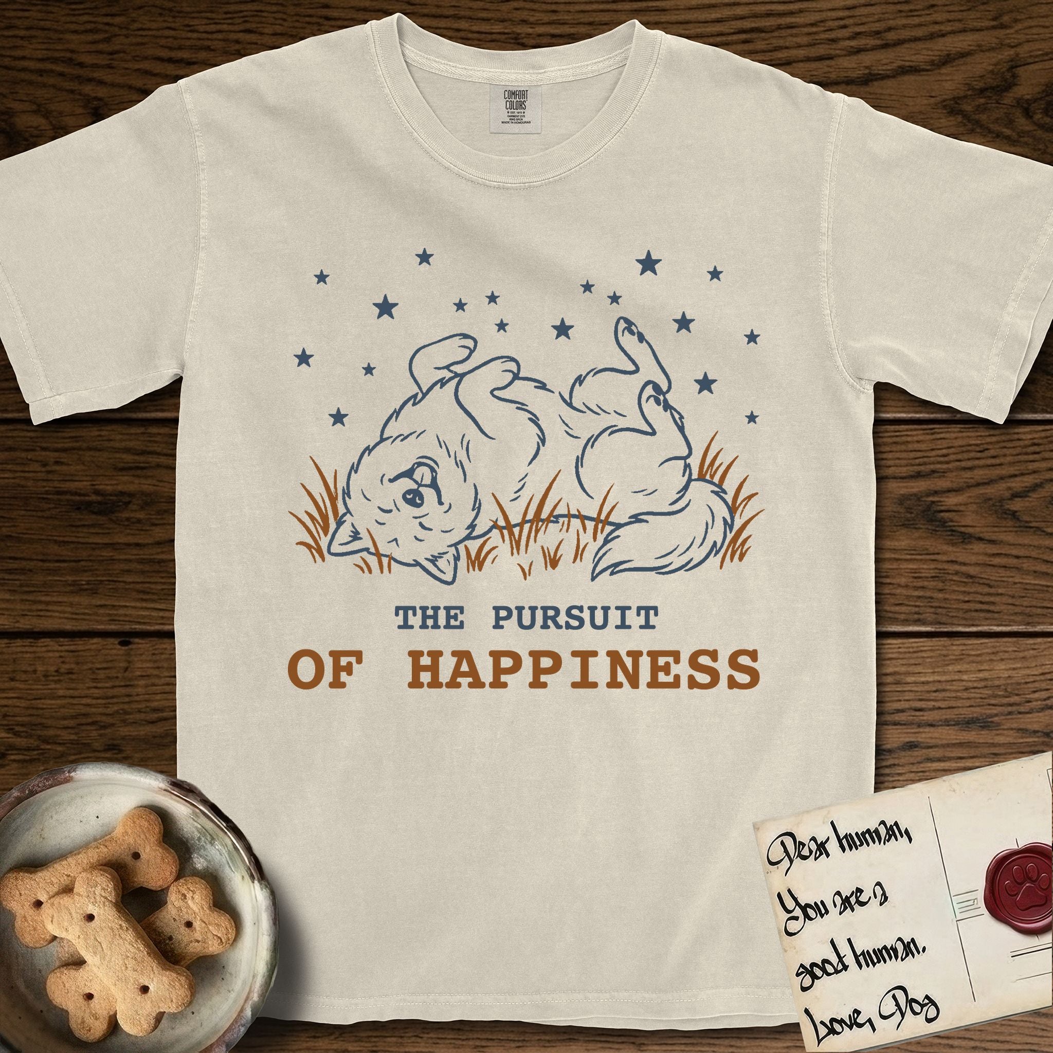 The Pursuit of Happiness T-Shirt flat lay in Ivory, featuring a faded vintage-Americana line-art Samoyed rolling on his back in the grass with paws in the air under a sky of stars, above “The Pursuit of Happiness” lettering.