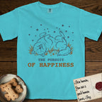 The Pursuit of Happiness T-Shirt flat lay in Lagoon Blue, featuring a faded vintage-Americana line-art Samoyed rolling on his back in the grass with paws in the air under a sky of stars, above “The Pursuit of Happiness” lettering.