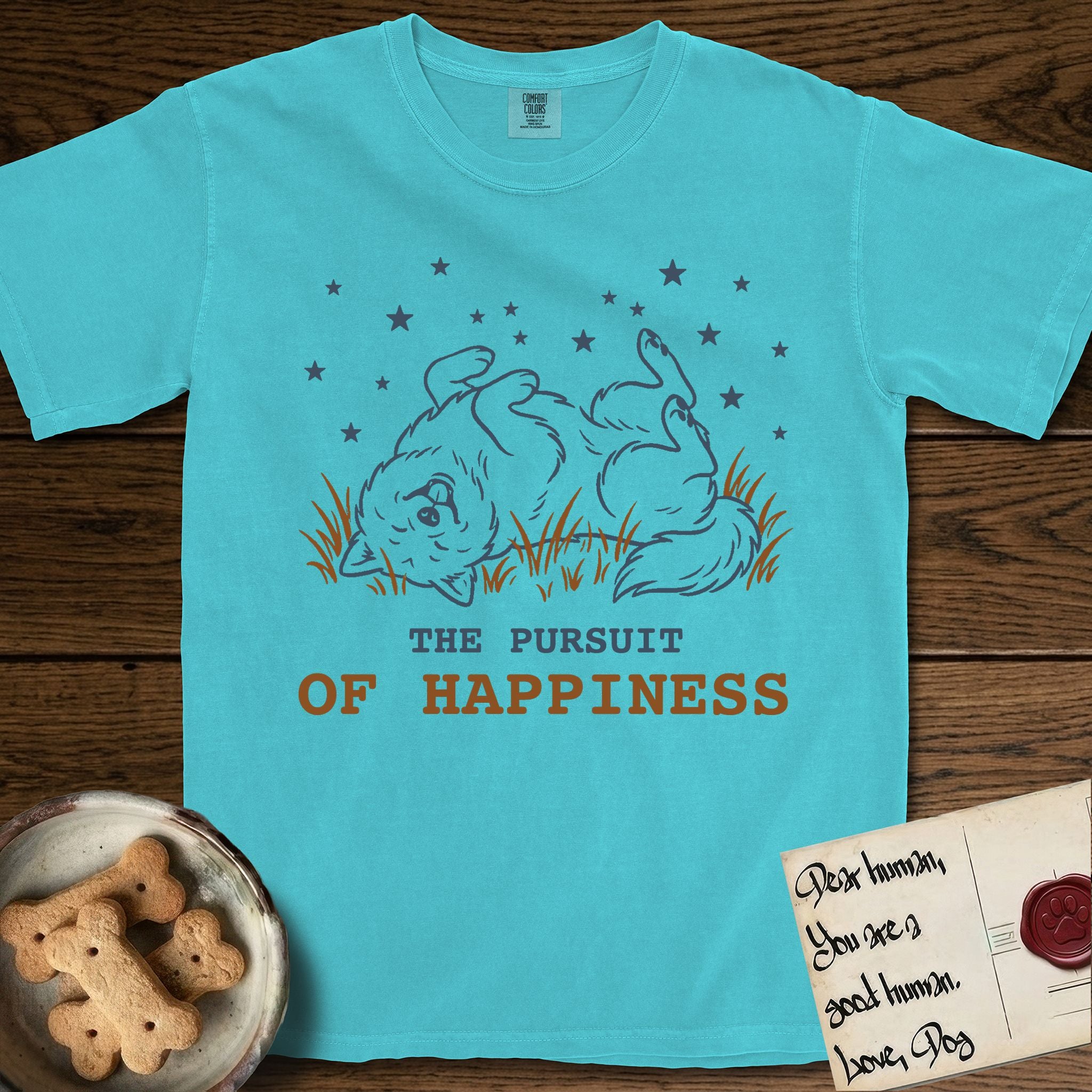 The Pursuit of Happiness T-Shirt flat lay in Lagoon Blue, featuring a faded vintage-Americana line-art Samoyed rolling on his back in the grass with paws in the air under a sky of stars, above “The Pursuit of Happiness” lettering.