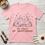 The Pursuit of Happiness T-Shirt flat lay in Light Pink, featuring a faded vintage-Americana line-art Samoyed rolling on his back in the grass with paws in the air under a sky of stars, above “The Pursuit of Happiness” lettering.