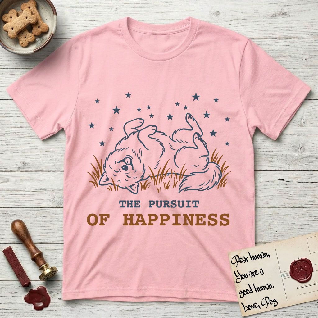 The Pursuit of Happiness T-Shirt flat lay in Light Pink, featuring a faded vintage-Americana line-art Samoyed rolling on his back in the grass with paws in the air under a sky of stars, above “The Pursuit of Happiness” lettering.