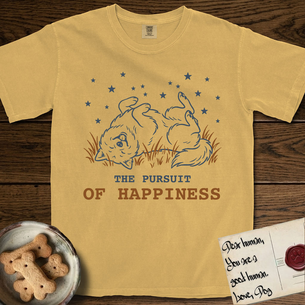 The Pursuit of Happiness T-Shirt flat lay in Mustard, featuring a faded vintage-Americana line-art Samoyed rolling on his back in the grass with paws in the air under a sky of stars, above “The Pursuit of Happiness” lettering.
