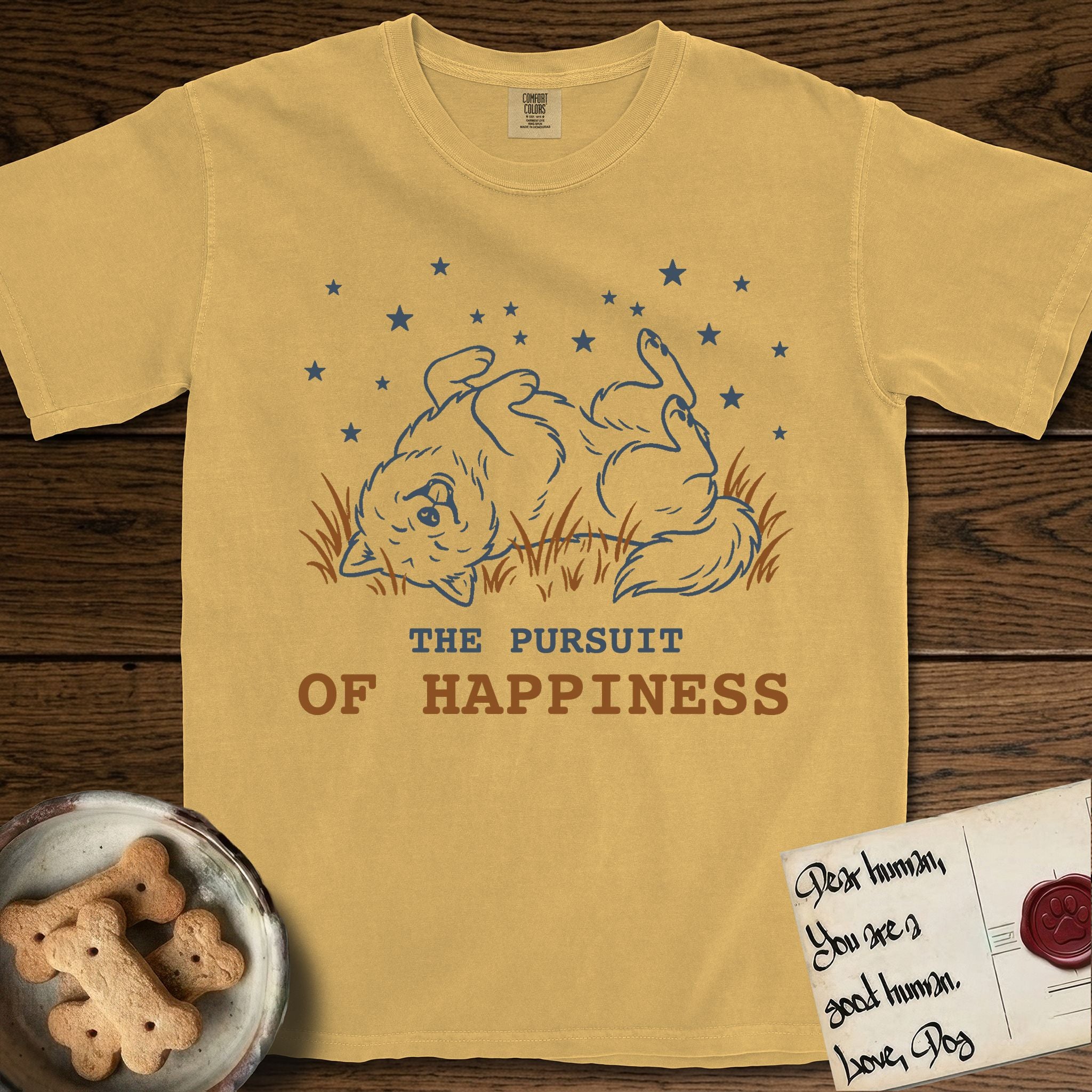The Pursuit of Happiness T-Shirt flat lay in Mustard, featuring a faded vintage-Americana line-art Samoyed rolling on his back in the grass with paws in the air under a sky of stars, above “The Pursuit of Happiness” lettering.