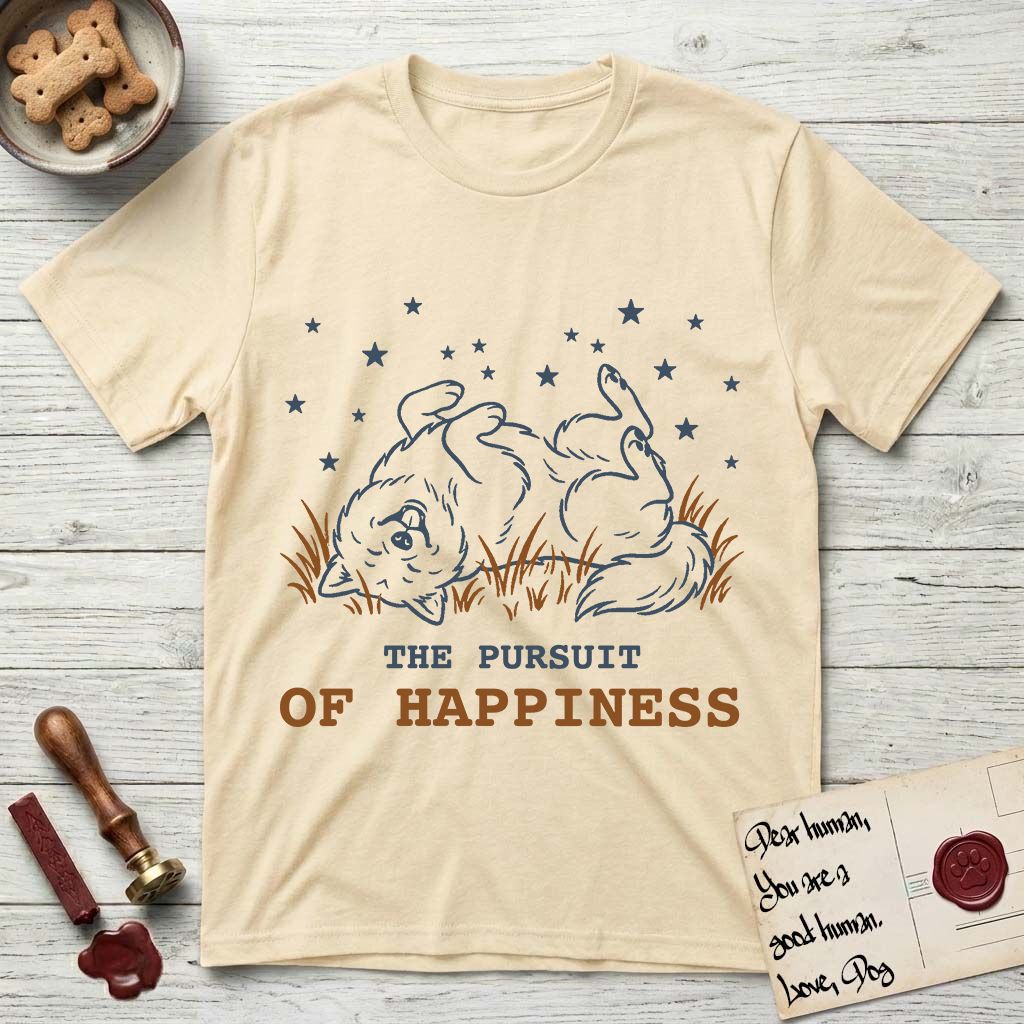 The Pursuit of Happiness T-Shirt flat lay in Natural, featuring a faded vintage-Americana line-art Samoyed rolling on his back in the grass with paws in the air under a sky of stars, above “The Pursuit of Happiness” lettering.