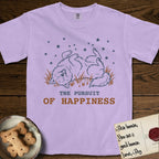 The Pursuit of Happiness T-Shirt flat lay in Orchid, featuring a faded vintage-Americana line-art Samoyed rolling on his back in the grass with paws in the air under a sky of stars, above “The Pursuit of Happiness” lettering.