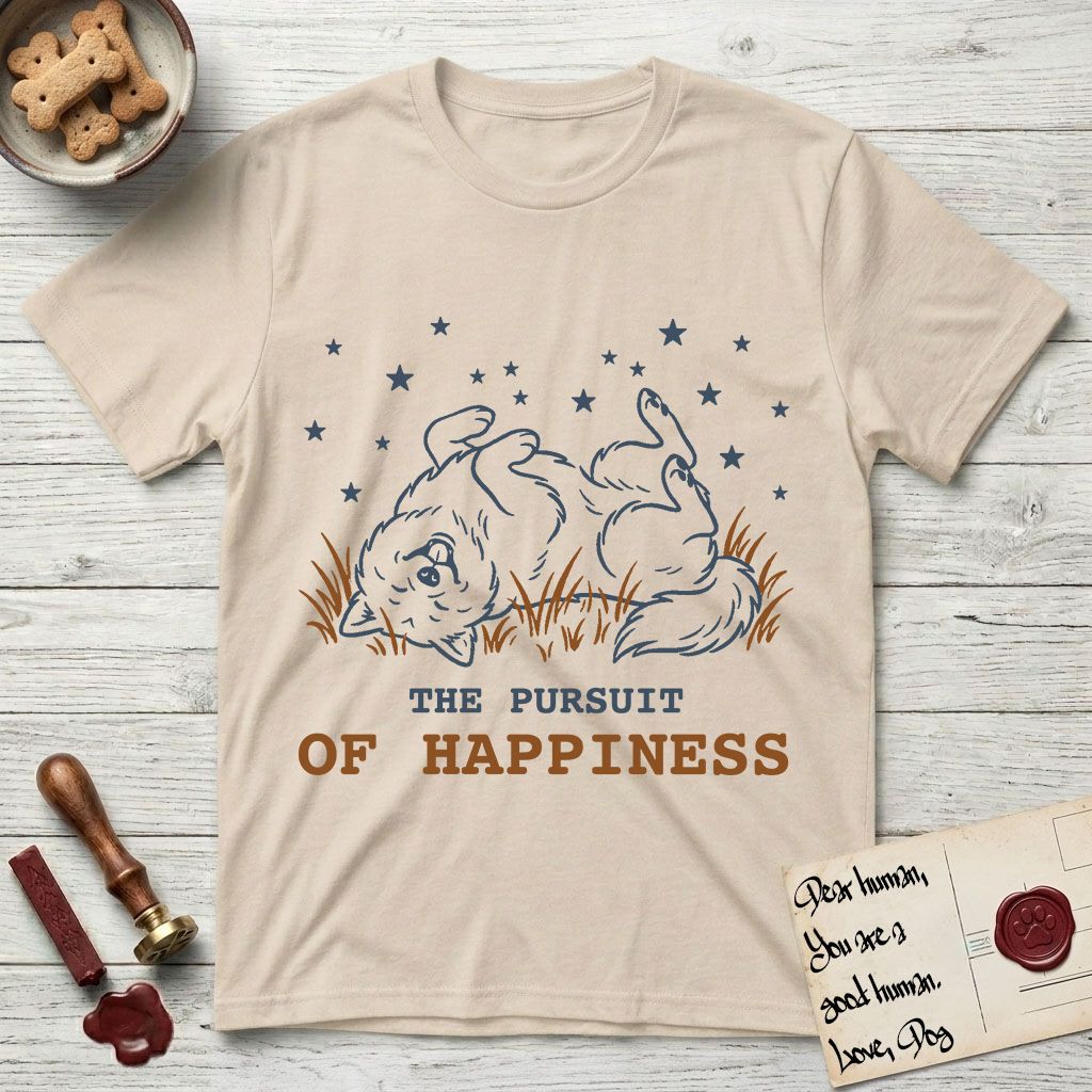 The Pursuit of Happiness T-Shirt flat lay in Sand, featuring a faded vintage-Americana line-art Samoyed rolling on his back in the grass with paws in the air under a sky of stars, above “The Pursuit of Happiness” lettering.