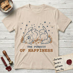 The Pursuit of Happiness T-Shirt flat lay in Sand, featuring a faded vintage-Americana line-art Samoyed rolling on his back in the grass with paws in the air under a sky of stars, above “The Pursuit of Happiness” lettering.