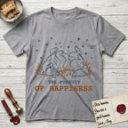The Pursuit of Happiness T-Shirt flat lay in Sport Grey, featuring a faded vintage-Americana line-art Samoyed rolling on his back in the grass with paws in the air under a sky of stars, above “The Pursuit of Happiness” lettering.